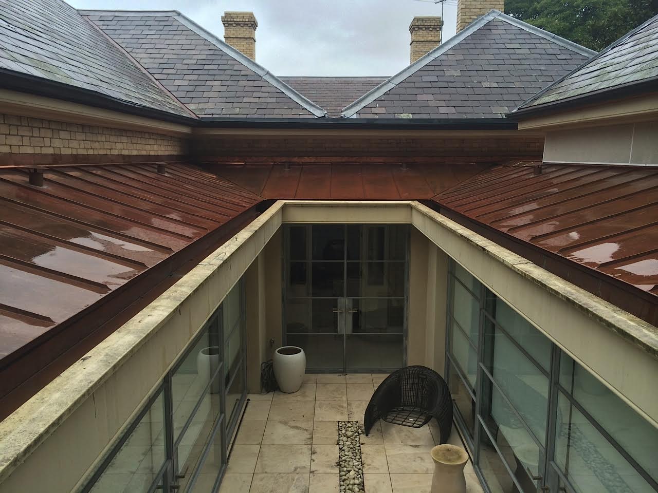 Modern building’s enclosed patio with glass doors, potted plants, metal chair, and copper zinc roofing system.