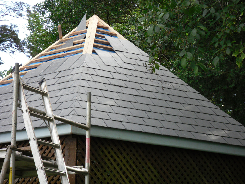 Pyramid-shaped slate roof under construction, grey shingles, wooden supports, and a metal ladder in view.