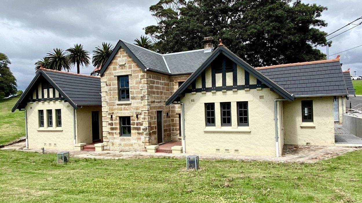 Single-storey house with stone and cream walls, dark roof tiles, gables, and elegant guttering on grassy lot.