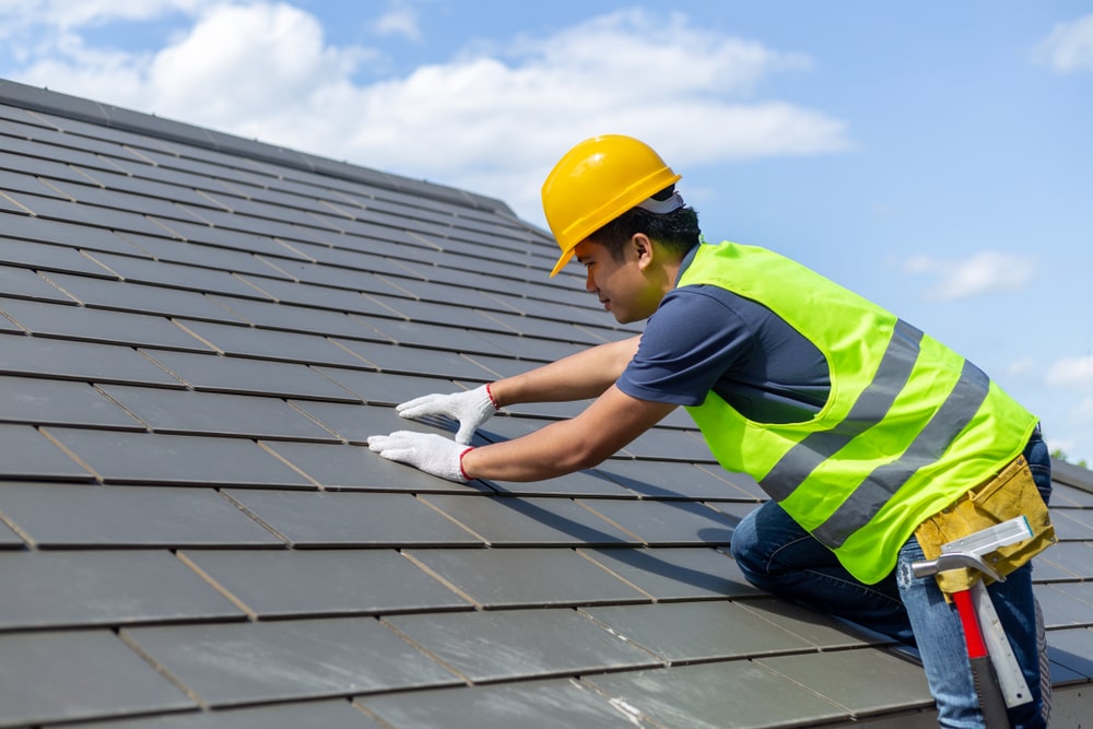A construction worker in a yellow hard hat installs tiles during slate roof repairs on a sloped roof.